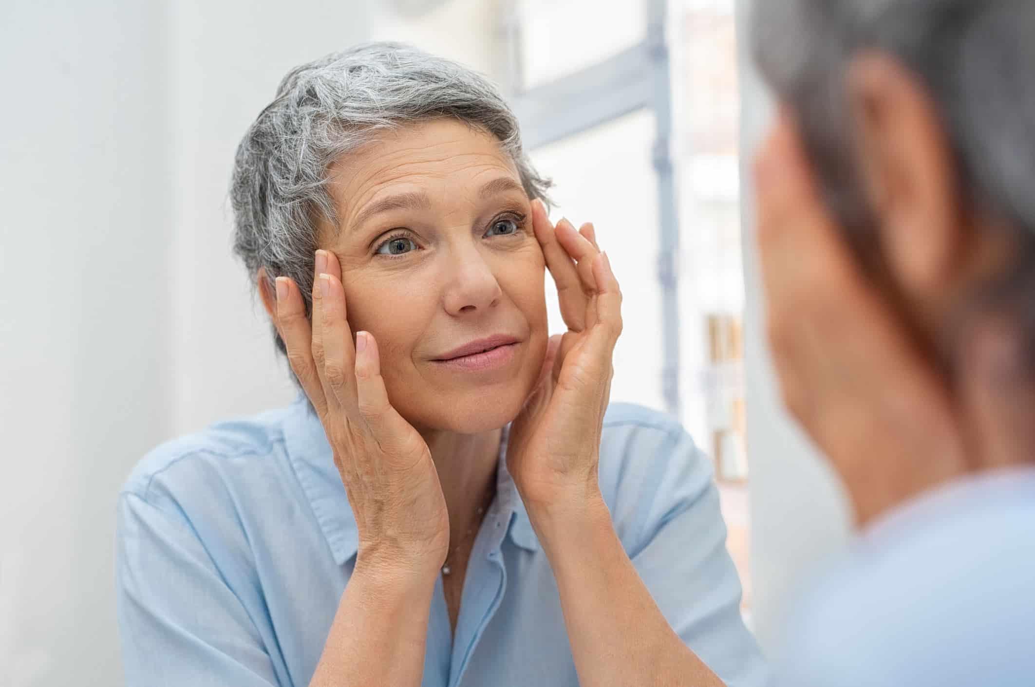 juvederm atlanta, image of a middle age woman with short gray hair gently touches and examines her face in the mirror.