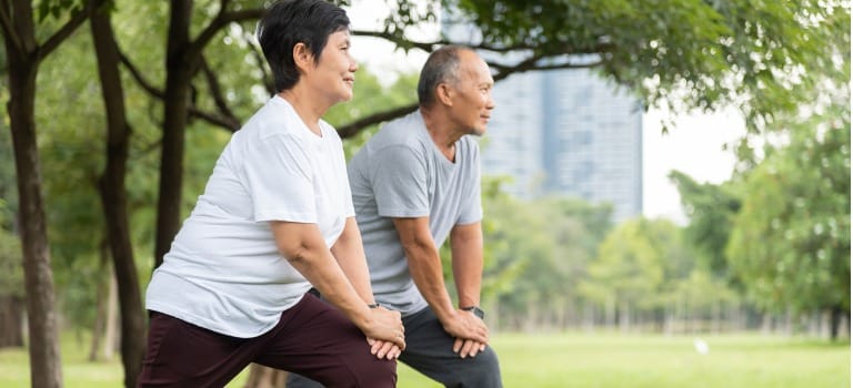 Knee Pain Specialists Woman wearing a white thsirt stretching with her left leg extended in front of her, knee bent, both hands on left knee. Man wearing a grey tshirt to her left is doing the same pose.