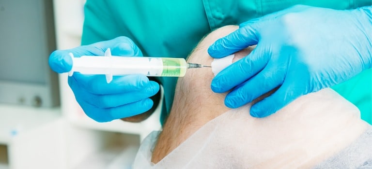 Knee Pain Specialists Medical practitioner wearing blue green scrubs and light blue medical gloves inserting a needle into a patient's knee