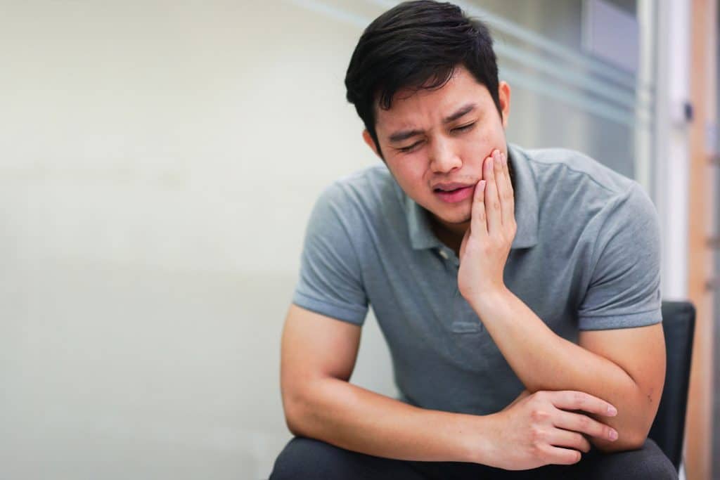 joint pain treatment Man wearing grey collared tshirt sitting with his elbows on his knees, grasping the left side of his face with his left hand