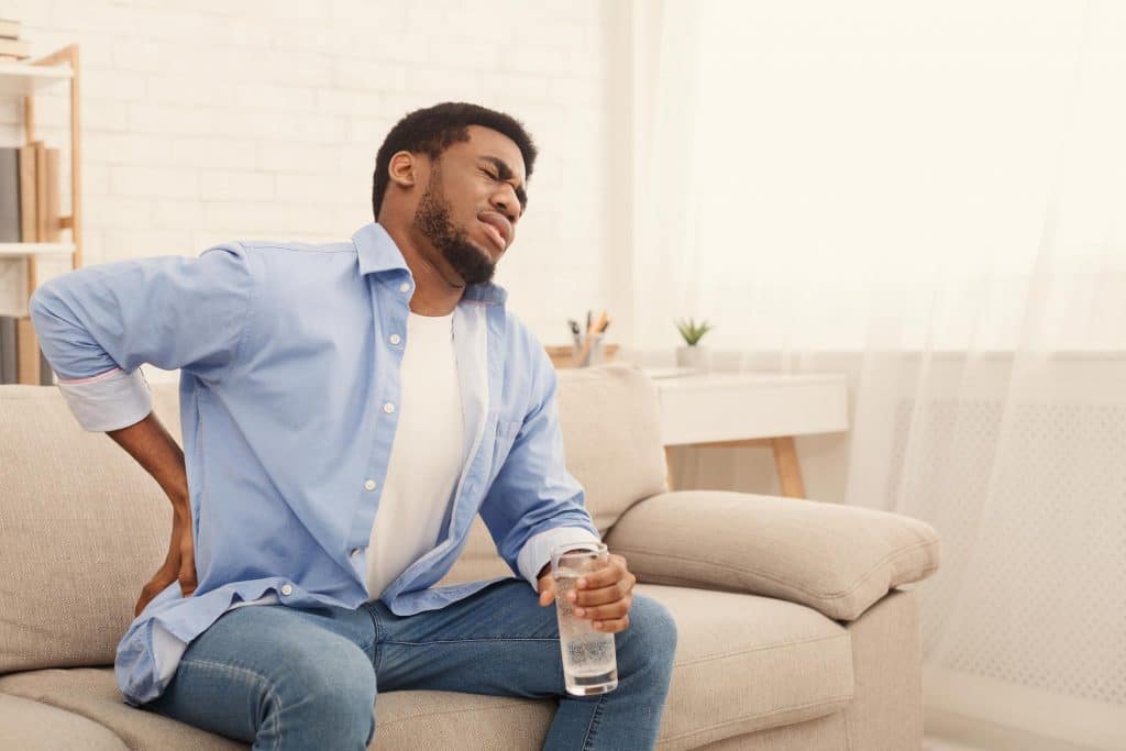 bone on bone pain man sitting on cream colored couch in a blue business shirt, clutching his lower back in his right hand and holding a glass of water in his left and grimacing