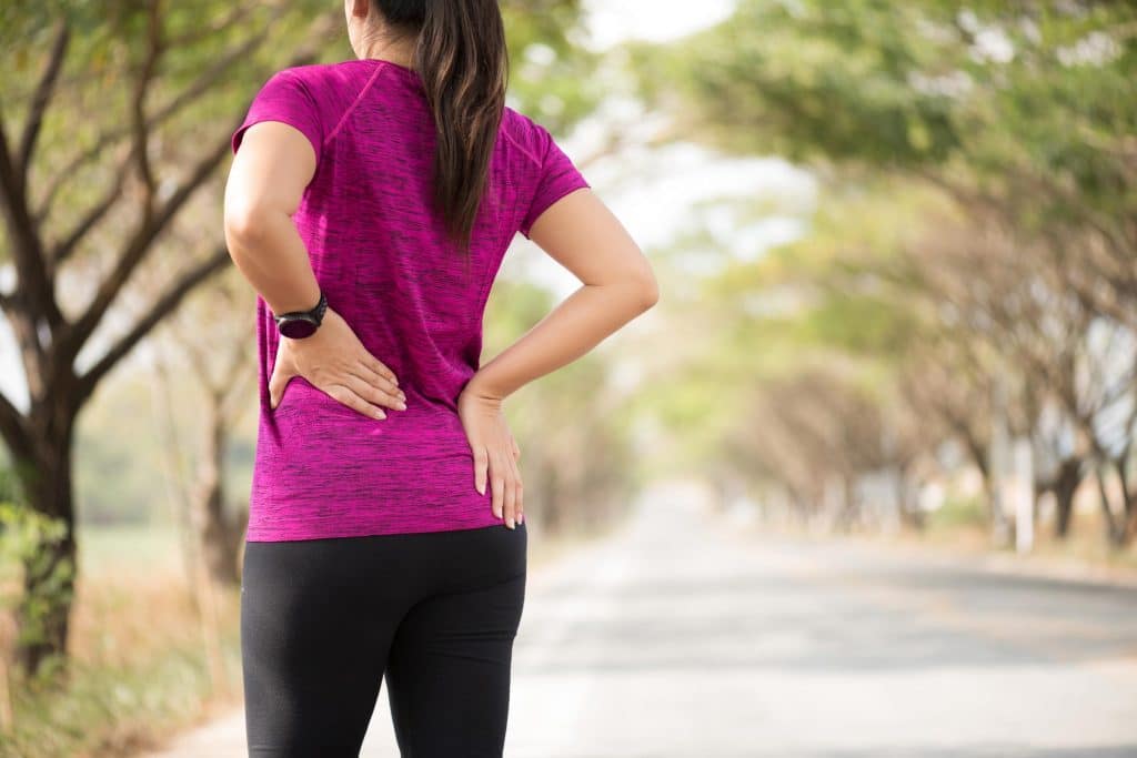 joint pain treatment Woman jogger wearing a magenta tshirt and black leggings standing and grasping lower back with both her hands