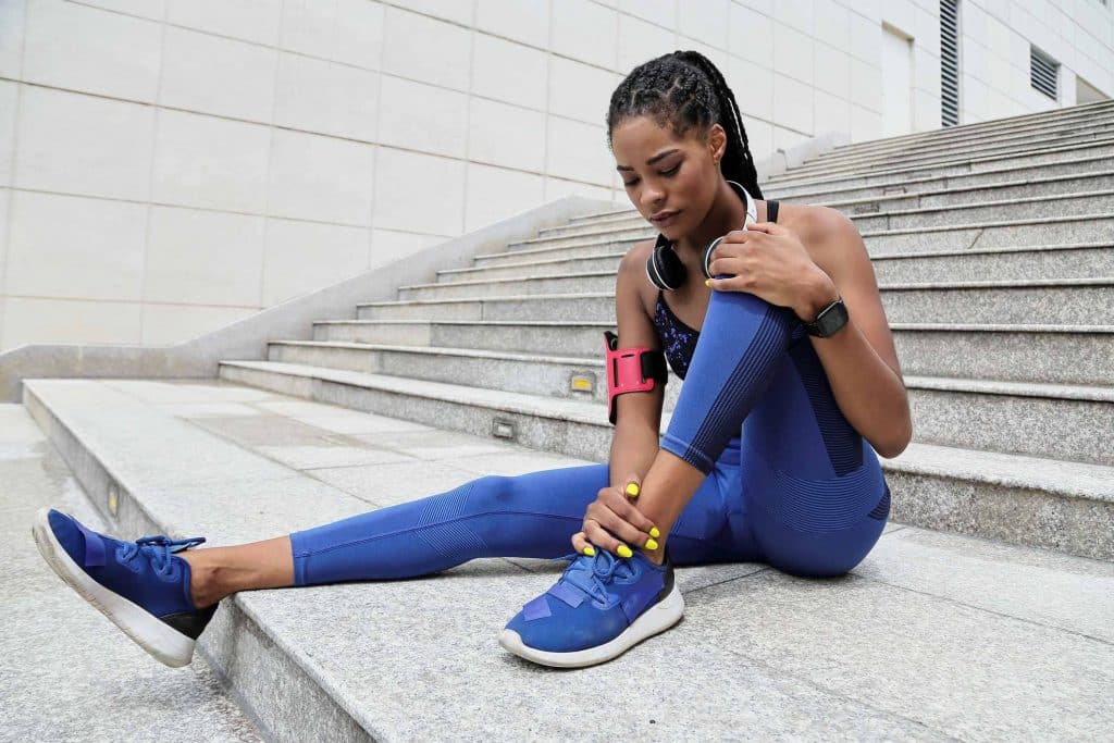 joint pain treatment Woman jogger wearing a black tank top, blue leggings and blue sneakers sitting on the ground with her right leg stretched out, and her left leg bent. Her right hand grasps her left ankle and her right hand rests on her left knee