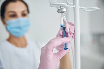 myers cocktail, image of a nurse injecting fluid into an IV drip with a female patient wearing a mask stands in the background.