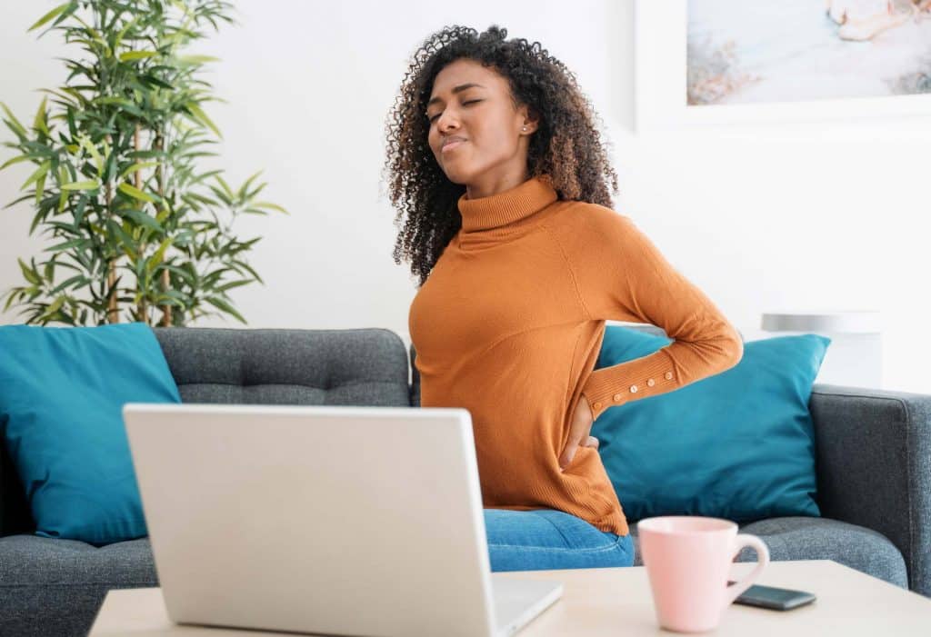 joint pain treatment Woman wearing an orange longsleeve shirt, sitting on a gray couch with blue pillows, working on her laptop at the coffee table, grasping her lower back with both hands with a look of pain