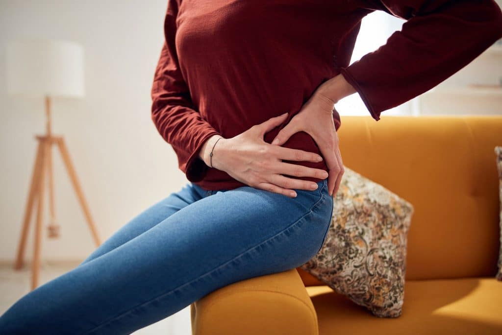 joint pain treatment Woman sitting on the arm of a gold yellow couch grasping her left hip with both her hands