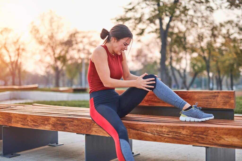 bone on bone pain woman sitting on a modern wood bench in multicolor leggings and a red tank top, holding her outstreched left knee in both hands
