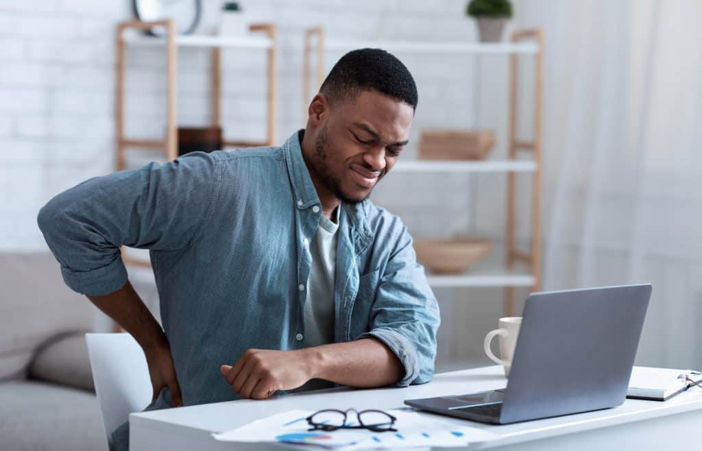 back pain specialists & doctors young man sitting at a computer desk in a blue business shirt, grimacing and holding a hand to his lower back
