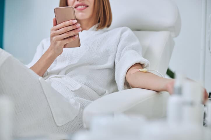 ozone therapy atlanta, image of a woman with short brown hair, sitting in a reclining doctor’s office chair, wearing a white robe, looking at her phone receiving IV treatment.