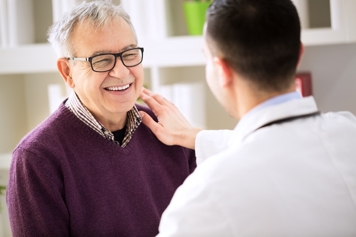 back pain specialists & doctors man with gray hair, glasses, and a maroon sweater smiling at a man in a white lab coat who is touching his shoulder as though sharing good news