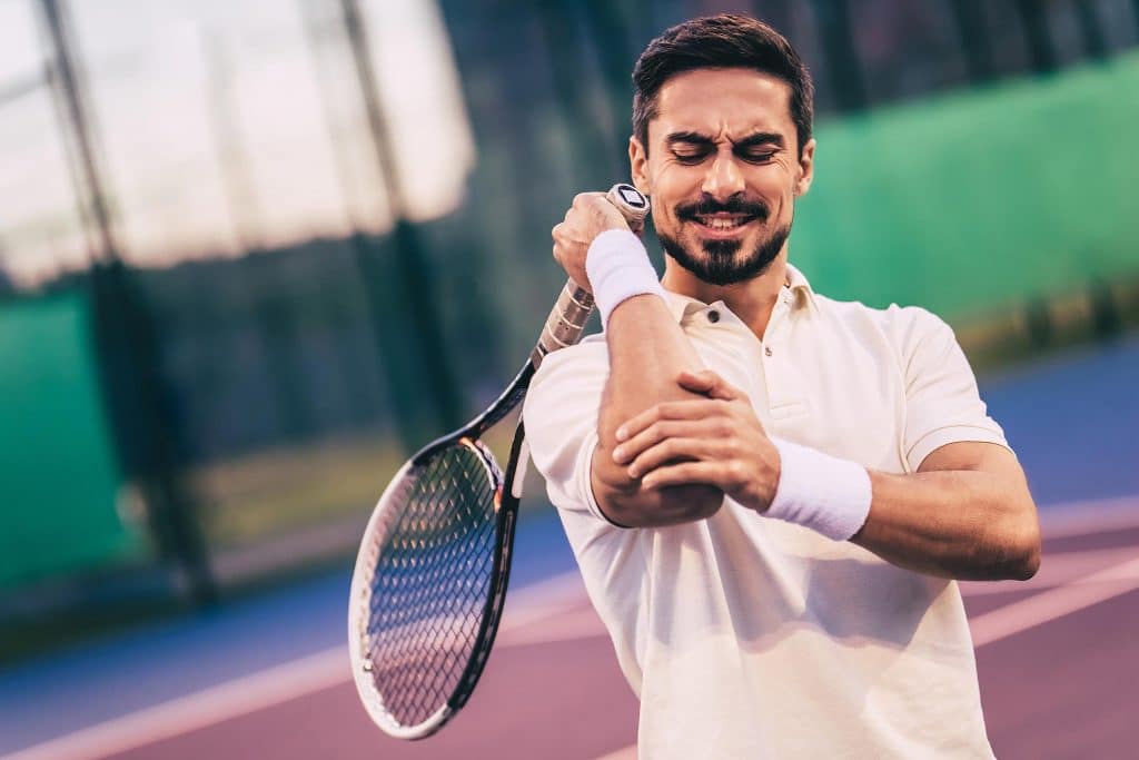 joint pain treatment Man tennis player wearing a white tee shirt clutching his right elbow with his left hand, tennis racket is in his right hand