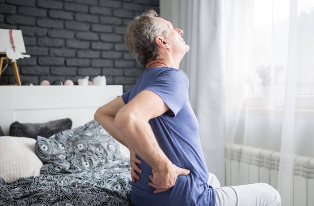 back pain specialists & doctors man with gray hair sitting on the edge of a bed in a purple shirt and gray sweatpants, holding both hands to his lower back in pain