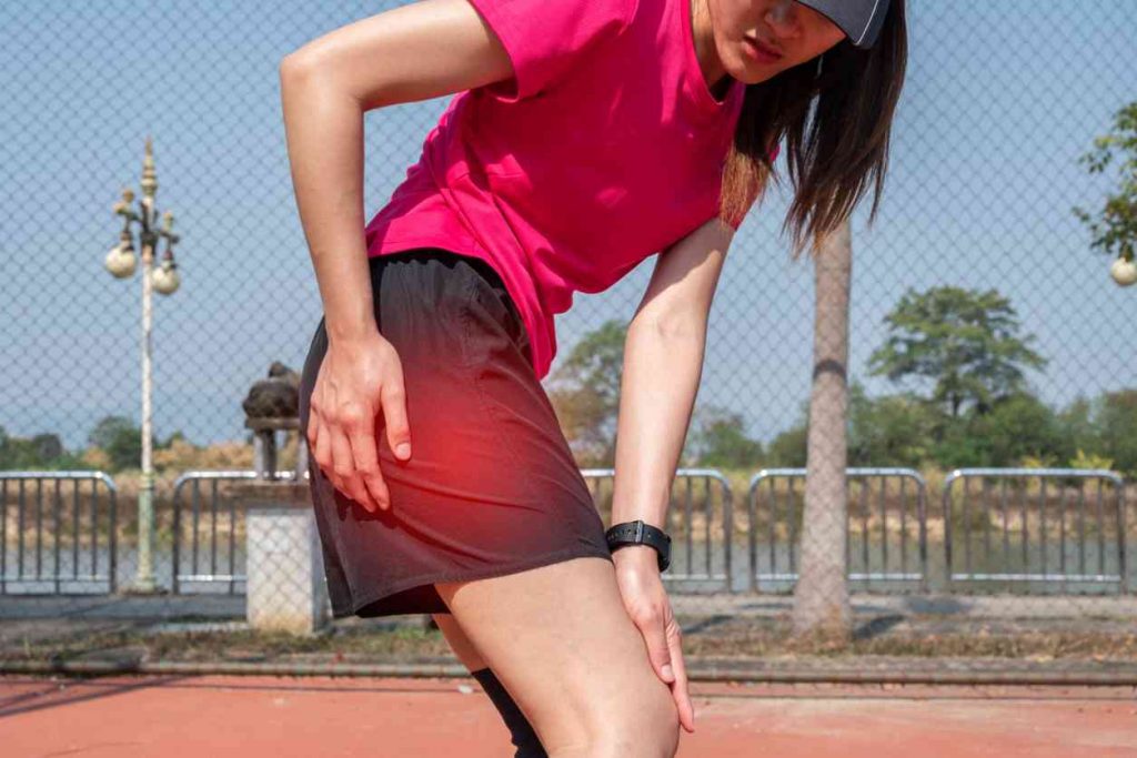Hip Pain Specialist Woman athlete standing, hunched over with her right hand grasping her right hip and her left hand on her right knee