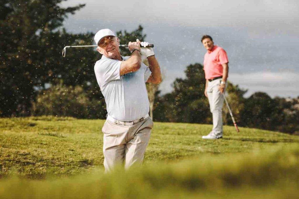 Hip Pain Specialist Older man with white collared tshirt swinging golf club and fellow player wearing a pink collared tshirt watching in the background