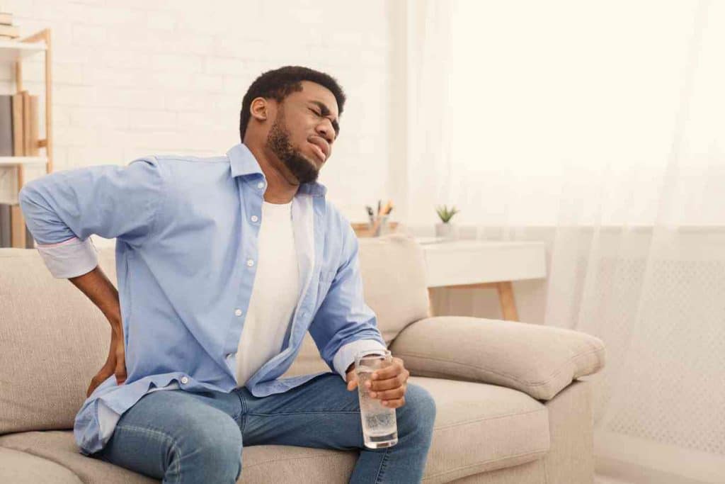 Hip Pain Specialist man sitting on cream colored couch in a blue business shirt, clutching his lower back in his right hand and holding a glass of water in his left and grimacing