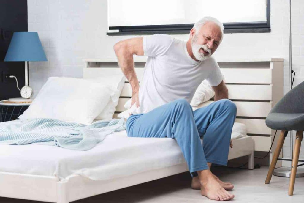 Hip Pain Specialist Elderly man sitting on edge of the bed hunched over with right hand on his lower back