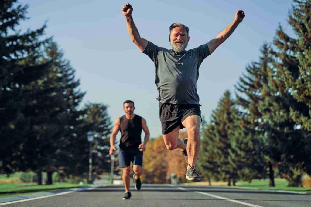 Hip Pain Specialist Older man wearing dark green tshirt and black shorts in mid-leap while running. Man wearing a black tank top and dark blue shorts running behind the first man.