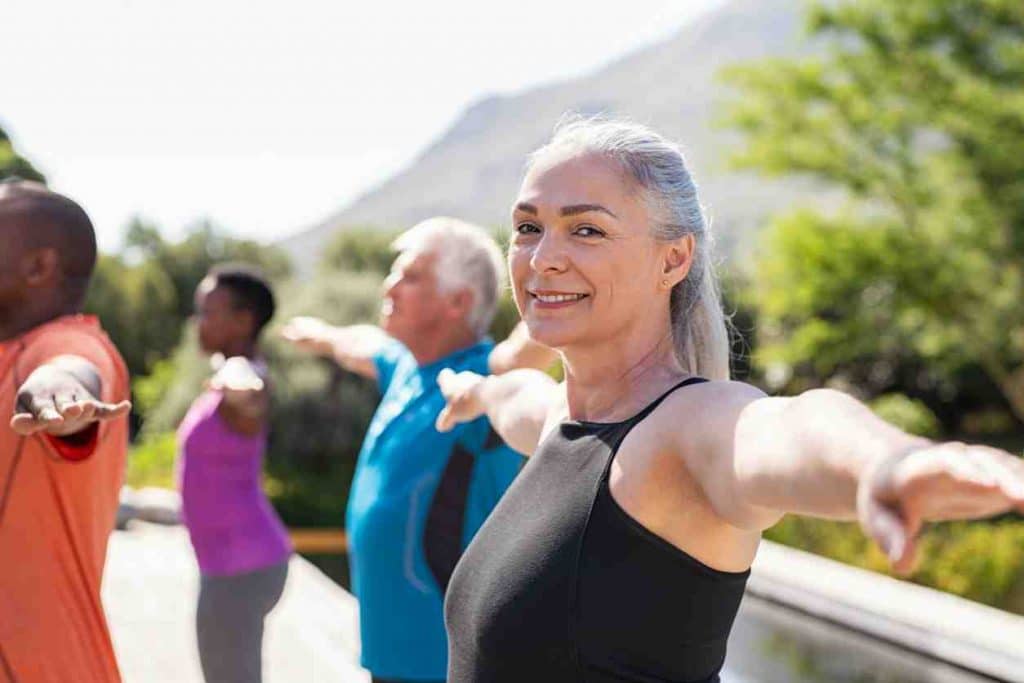 Hip Pain Specialist Woman with gray hair smiling while stretching her arms out from each side in a yoga pose. Others around her doing the same yoga pose