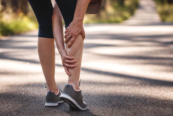 Trigger Point Injections, Woman jogger wearing black capri leggings and dark gray sneakers is bent over grasping her right calf muscle with both of her hands