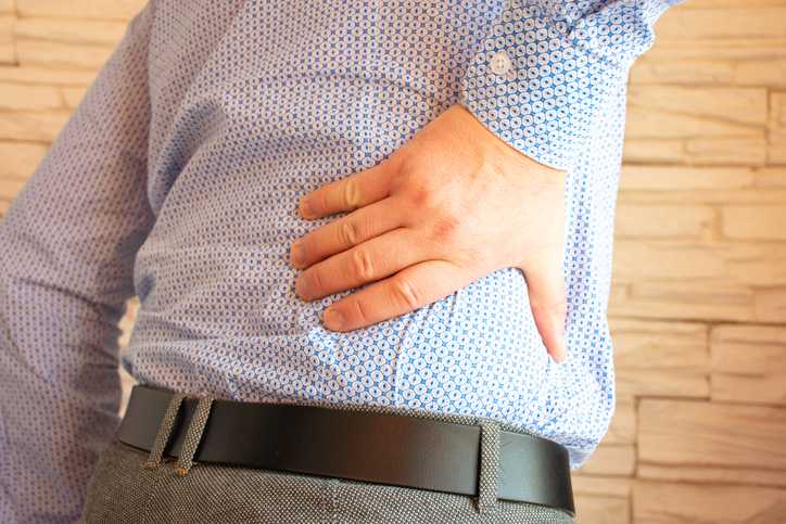 spinal stenosis treatment, image of a man, dressed in professional clothing, grasp his lower back with his right hand.