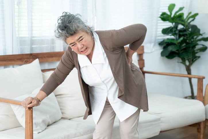 spinal stenosis treatment, image of an elderly woman leaning forward to grasp her lower back with her left hand, while her right hand grasps the arm of a couch for support.