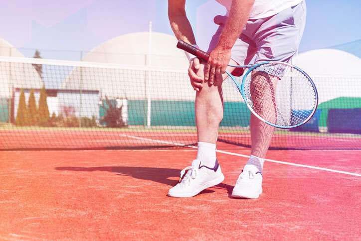 Male tennis player bends over clutching his right knee with both of his hands while holding his tennis racket with his left hand.