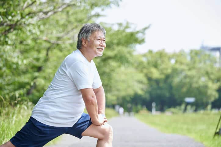 Prolozone Therapy, Man wearing white tshirt and navy shorts stretching on a paved path in a park with his left leg extended out, left knee bent and both his hands on his left knee