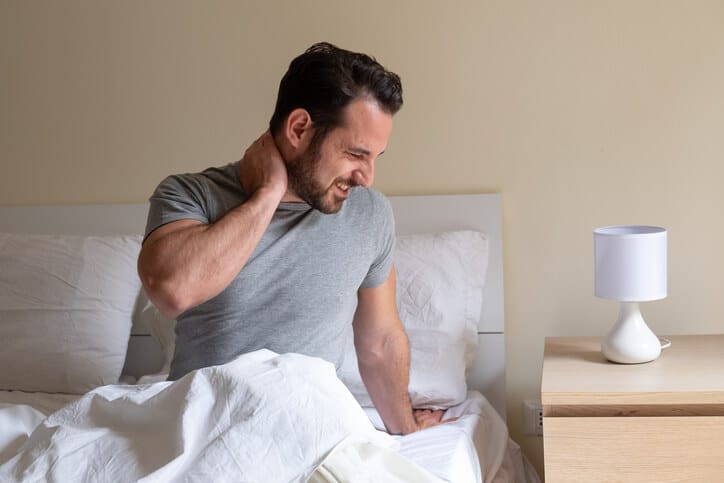 neck pain and headache relief Man wearing a grey tshirt sitting up in bed with his right hand grasping the back of his neck and a look of pain on his face