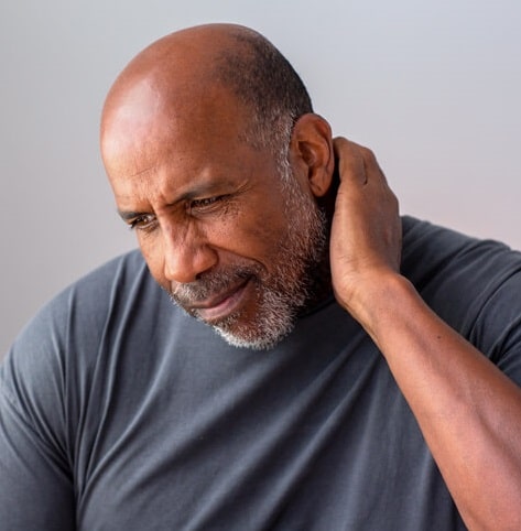 neck pain and headache relief Older man wearing a dark grey tshirt grasps the back of his neck with his left hand and has a look of pain on his face