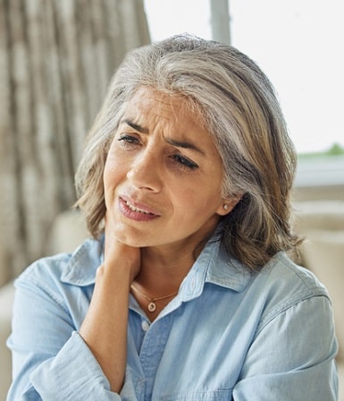 Prolozone Therapy, Woman with gray hair wearing a light blue collared shirt has a look of pain on her face and grasps her neck with her right hand