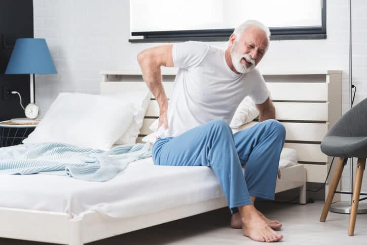 Prolotherapy, Old man with white hair and facial hair, wearing a white tshirt and blue pajama pants sits on the edge of the bed grabbing his lower back with his right hand