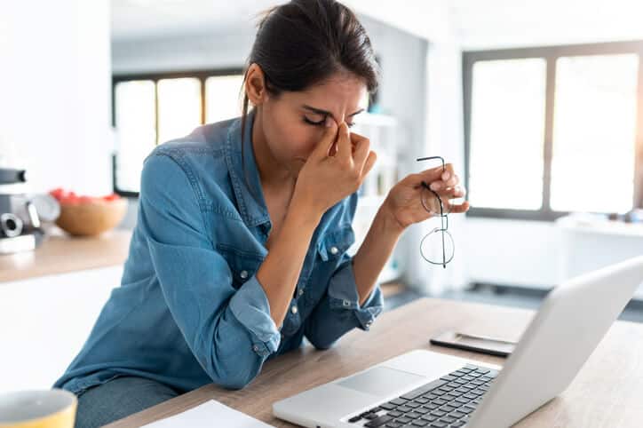 neck pain and headache relief Woman wearing a blue long sleeve shirt works on her laptop, elbows on the desk, left hand holding her eye glasses, right hand pinching the bridge of her nose