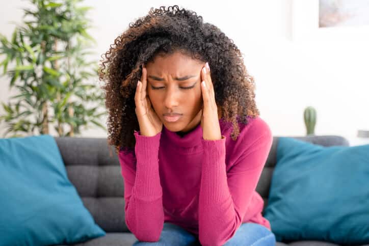 neck pain and headache relief Woman wearing a magenta sweater sitting on a grey couch with blue pillows holds the sides of her face with both hands, eyes closed, with a look of pain on her face
