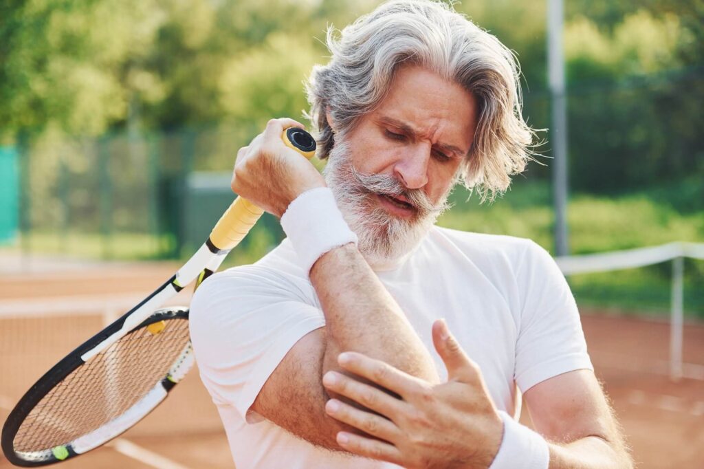 elbow and upper arm pain Man tennis player wearing a white tee shirt clutching his right elbow with his left hand, tennis racket is in his right hand