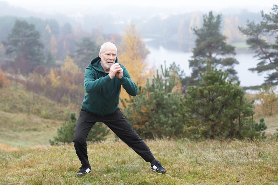 Prevent Orthopedic Surgery, Autumn portrait of sporty healthy mature male in hoodie and running shoes exercising outdoors, practicing side lunges. Elderly bearded man in sportswear warming up before morning run in park