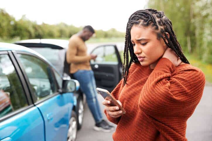 Auto Injury Treatment, Woman wearing orange sweater with concerned look on her face looking at her phone and standing next to a blue car. Man in background leans against his car, looking at his phone
