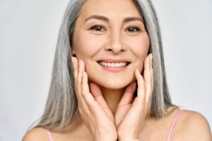 restylane treatment, image of a smiling woman, with long gray hair, gently touching her face with both her hands.