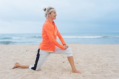 Regenerative Medicine, Woman wearing an orange zip-up sweatshirt and rolled-up gray sweatpants stretching on the beach on one knee, left leg extended out with both hands on her left leg