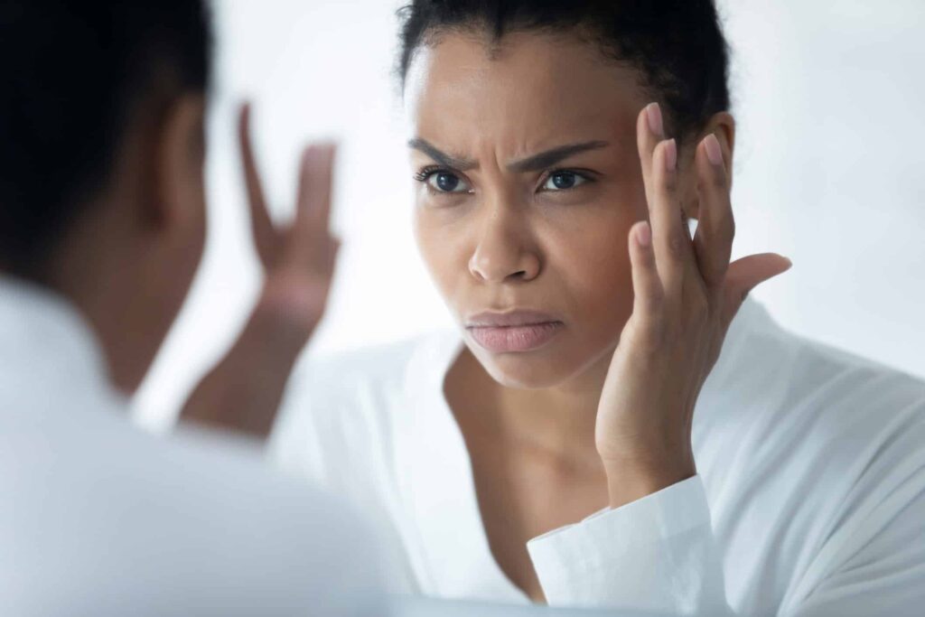 Restylane Filler​, image of a young woman with a frustrated expression looking at her face in the mirror and touching the right side of her face with her right hand.