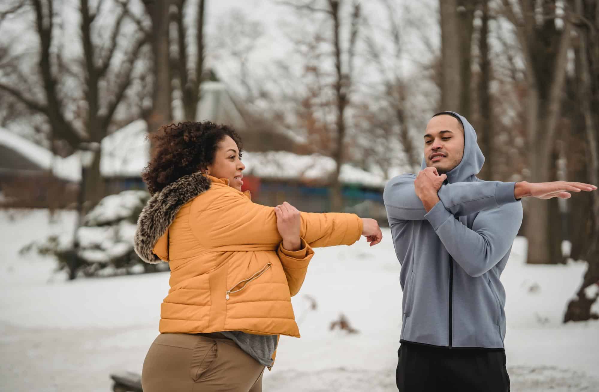 joint pain in cold weather, a woman wearing an orange winter coat and a man wearing a gray zip-up sweatshirt do arm stretches together in a snow covered park.