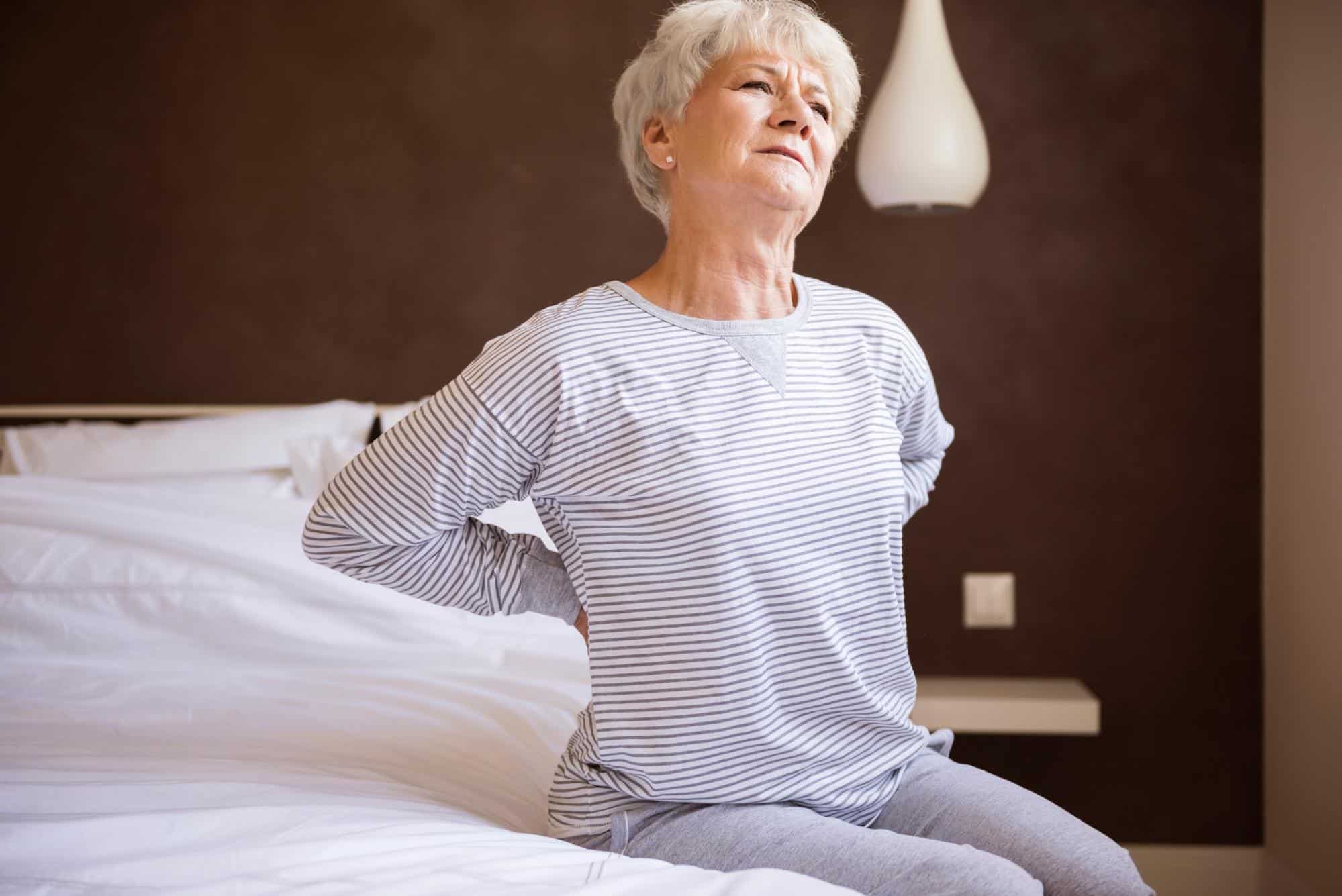 joint pain in cold weather, an older woman wearing a gray and white striped shirt and gray pants sits on the edge of her bed while she bends her back and has her hands placed on her lower back.