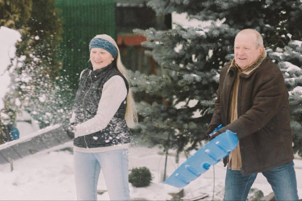joint pain in cold weather, a woman wearing a black winter vest, gray sweater, jeans and a teal knit headband and a man wearing a brown winter coat and jeans shovel snow together.