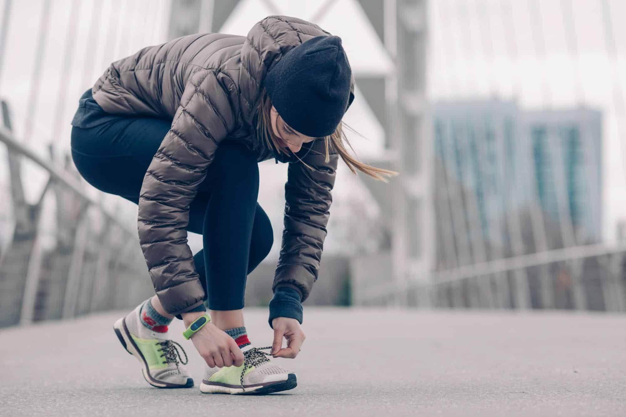 joint pain in cold weather, a woman jogger wearing winter athletic attire bends down to tie the sneaker on her right foot.