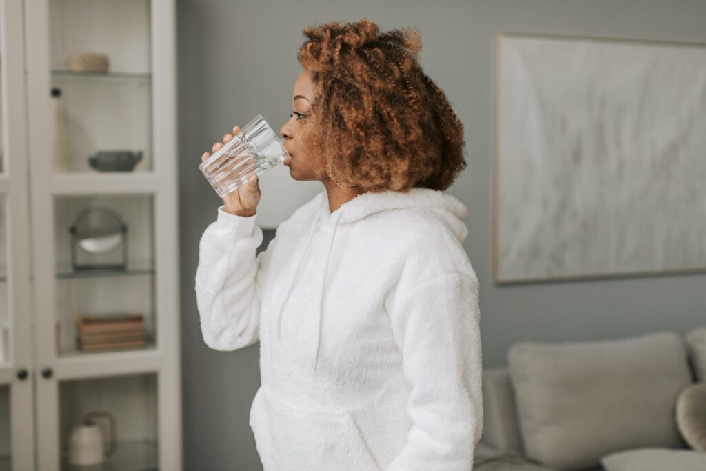joint pain in cold weather, a standing woman wearing a fuzzy white hooded sweatshirt drinks a glass of water.