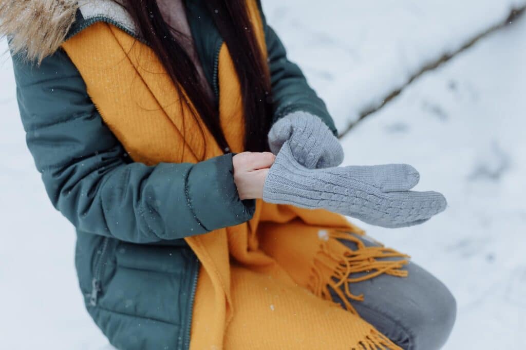 joint pain in cold weather, a sitting woman wearing a teal winter jacket and orange scarf places a knit gray mitten on her right hand.