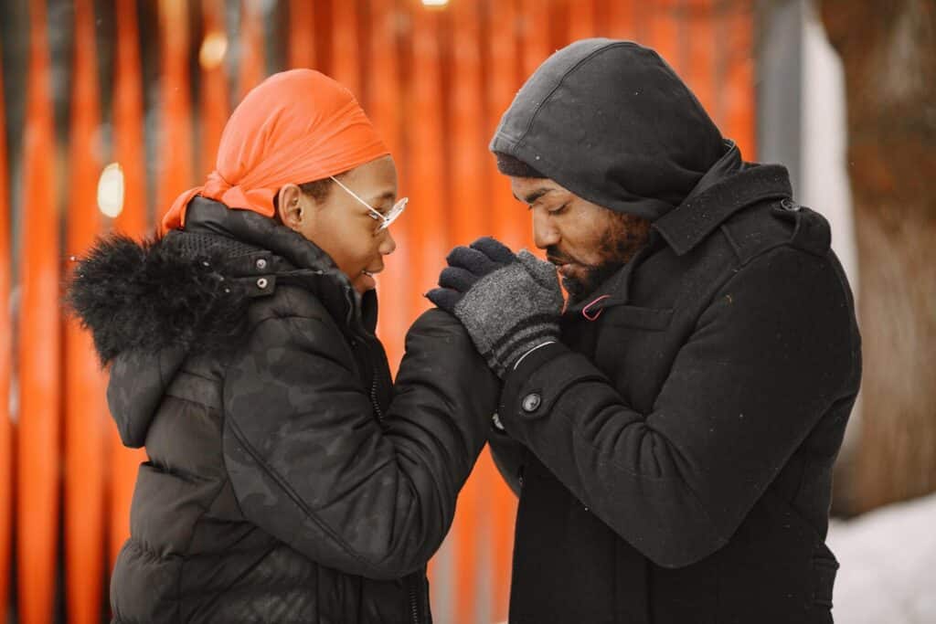 Cold weather injuries, Man and woman wearing winter jackets and gloves stand outside together while the man grasps the woman’s hands and blows on them to keep them warm.