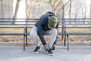 Cold weather injuries, Man wearing a navy winter jacket, gray jogger pants, a green knit cap and black sneakers sits on a park bench to tie the shoe on his left foot.