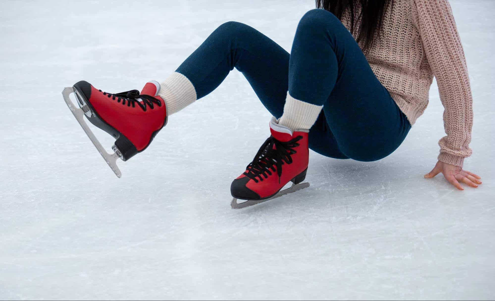 Cold weather injuries, Woman with long dark hair wearing a beige sweater, navy leggings and red ice skates picks herself up after falling on the ice.