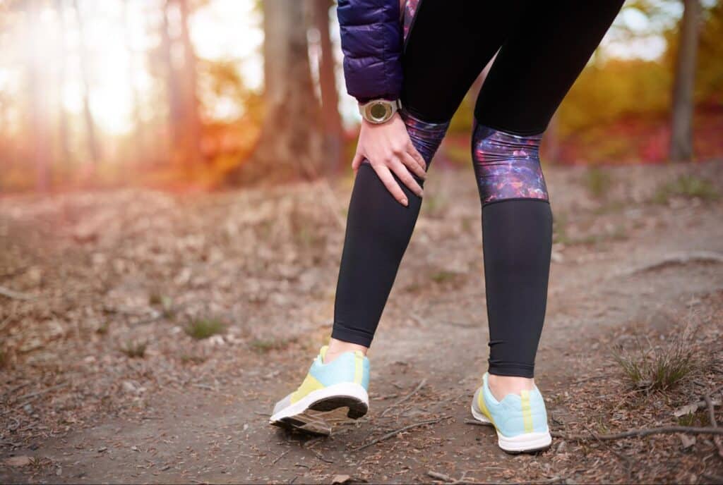 Cold weather injuries, Woman jogger stands in the middle of a forest dirt path and bends down to grasp her left knee with both hands.