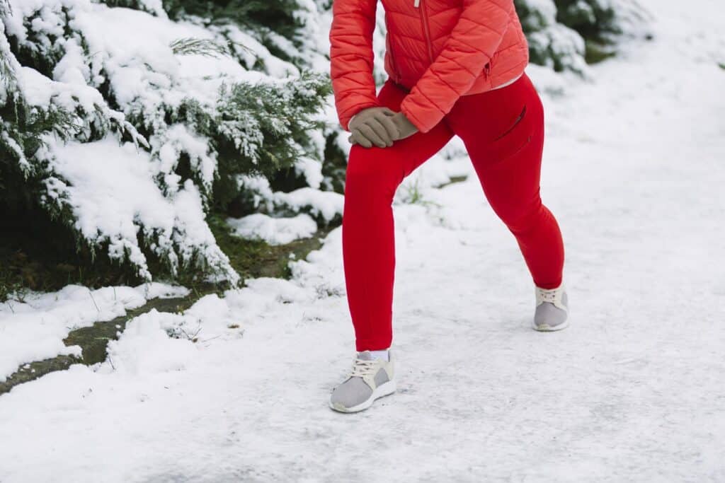 Cold weather injuries, Woman on a snowy path wearing red athletic leggings, coral puffy jacket and brown gloves stretches her legs with her right leg extended in front of her left leg extended behind her.